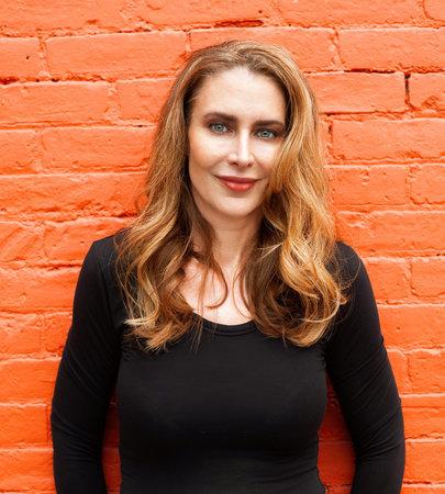 Photo of Abbott Kahler, a Caucasian woman with long reddish blonde hair and red lipstick wearing a long-sleeve black shirt standing against an vibrant orange brick wall