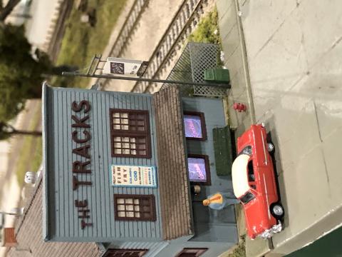 Part of a model train set, the image is a scaled model of "The Tracks" in Cary, Illinois, a blue-green shed building with its name on the front above windows and a door. A red 1950s Cadillac scaled model is parked in front of the model building.