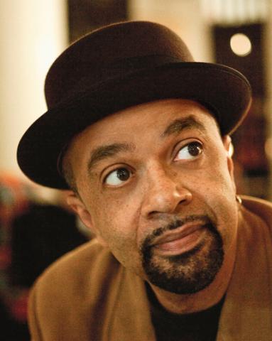 A headshot of James McBride, an African American male with dark facial hair and brown eyes wearing a fedora-style hat and light brown suit jacket looking candidly away from the camera. 