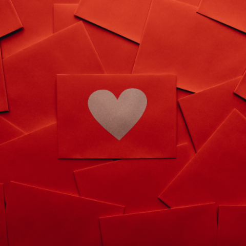 A red envelope with brown paper heart cut into it, on top of a pile of other red envelopes. 