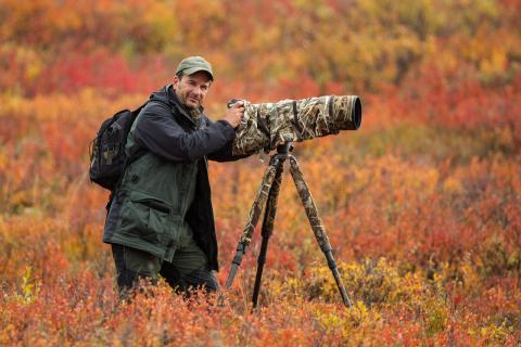 Stan Tekiela, a nature photographer with a camouflaged, heavy-duty camera standing on a tripod against a backdrop of fall foliage.