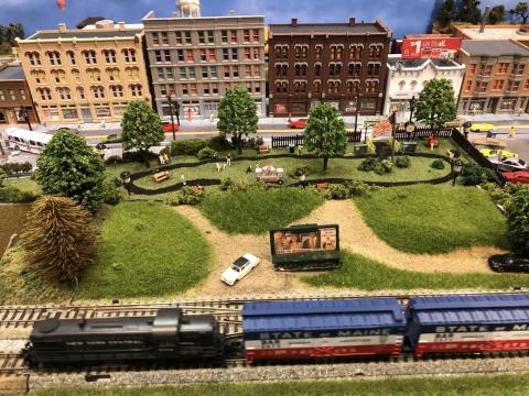 A model train and a landscape with green grass, trees, and small building are made to scale. The display is filled with detailed and carefully place objects. 