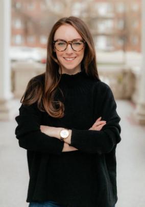 Barbara Ginty, a Caucasian female seemingly in her 30s with long brown hair, thick-rimmed black glasses and a full smile. Her arms are crossed across her chest and she is wearing a large, long black sweater. Background is a blurred cityscape.