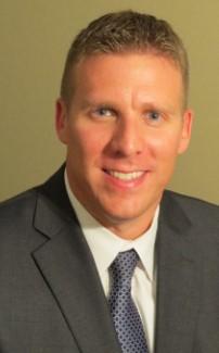 John Daly, a Caucasian male with short blonde hair, light eyes, and a full smile. He is wearing a gray suit, over a white dress shirt with a gray tie. Photo taken against a light olive green background. 