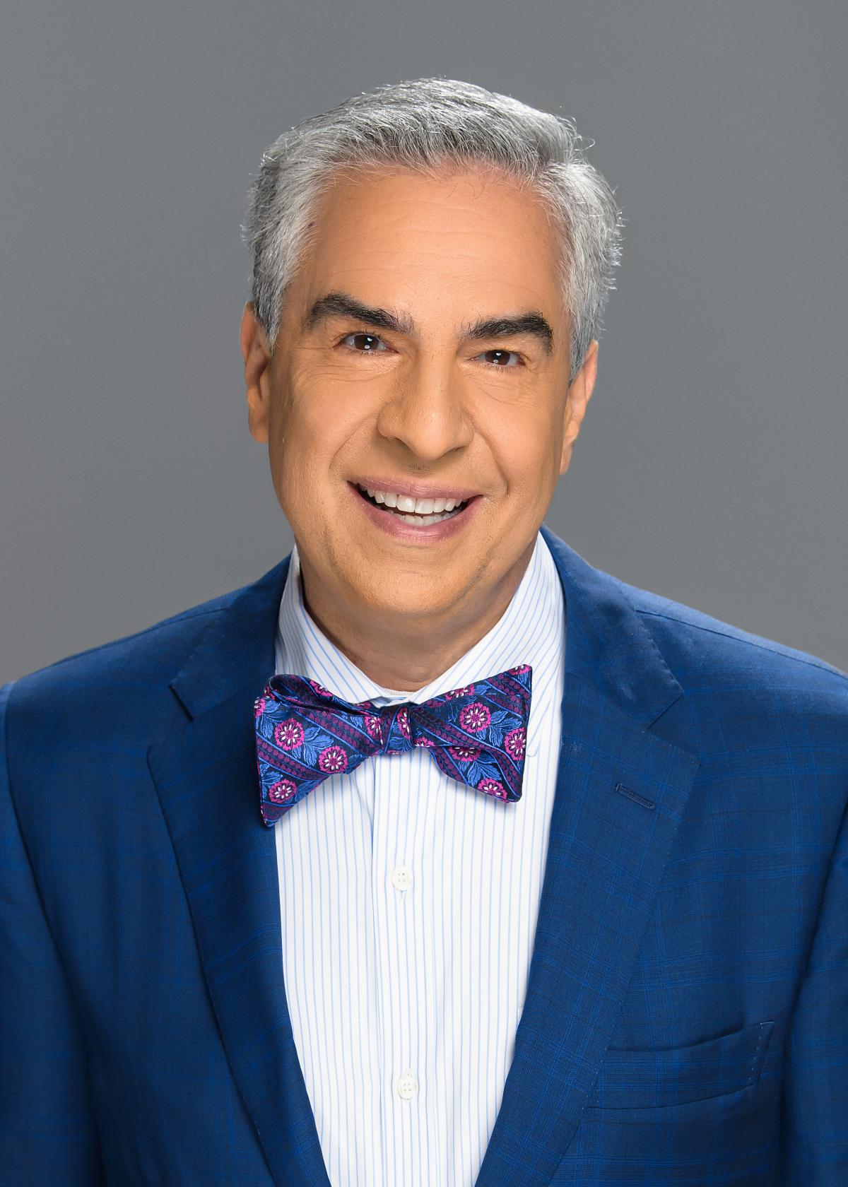 Paul Lisnek, an olive-skin male with gray and black hair and a large smile, wearing a rich blue suit jacket and matching paisley bow tie over a white button-up dress shirt.