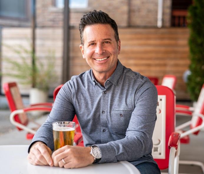 Steve Dolinsky, a caucasian male with medium length dark hair slicked back, wearing a gray button-up shirt and dark wash jeans. He is sitting in an outdoor diner of red and white metal chairs, with a draft pale ale on the table in front of him.